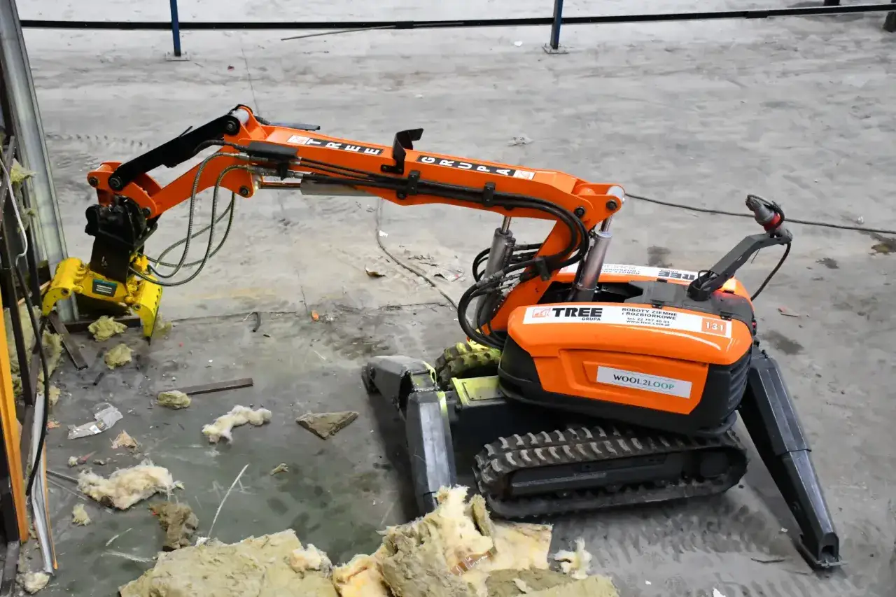 Pomarańczowe roboty wyburzeniowe z chwytakiem pracują na placu budowy, rozbijając materiały izolacyjne.