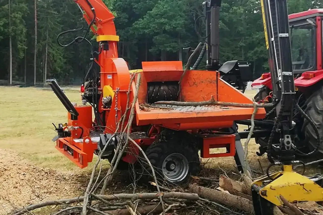 Pomarańczowa maszyna do zrębki pracuje, rozdrabniając gałęzie. W tle widać czerwony traktor i las.