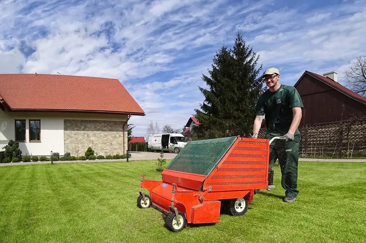 Mężczyzna w zielonym ubraniu pcha wertykulator, przygotowując trawnik do wiosennych prac. Idealny czas, kiedy wertykulować trawnik, to właśnie teraz!