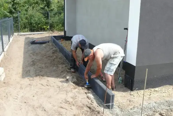 Dwóch mężczyzn układa krawężniki, jeden używa młotka do zagęszczania betonu.