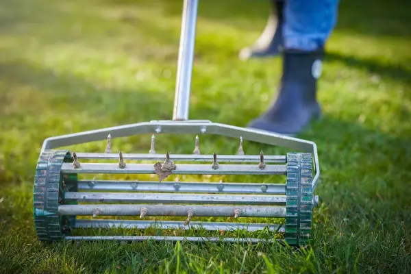 Aerator do trawy z kolcami na zielonej trawie. W tle widać nogi w gumowcach.